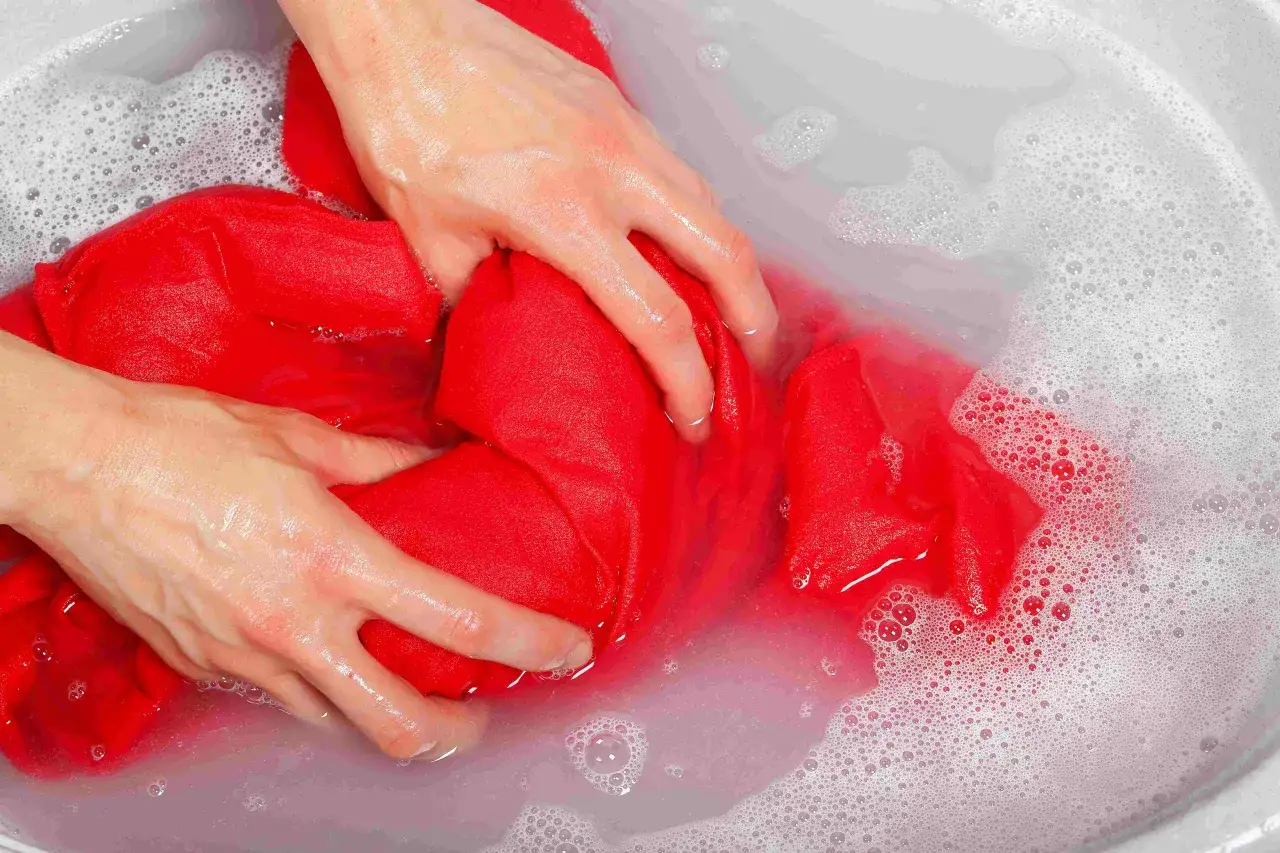 Mani che lavano un capo rosso in una bacinella con acqua e schiuma, per smacchiare con candeggina.