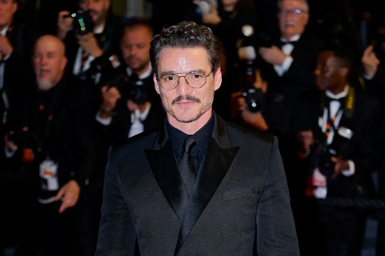 Pedro Pascal sfoggia un elegante outfit giacca nera uomo, con camicia e cravatta abbinate, occhiali da vista e baffi.