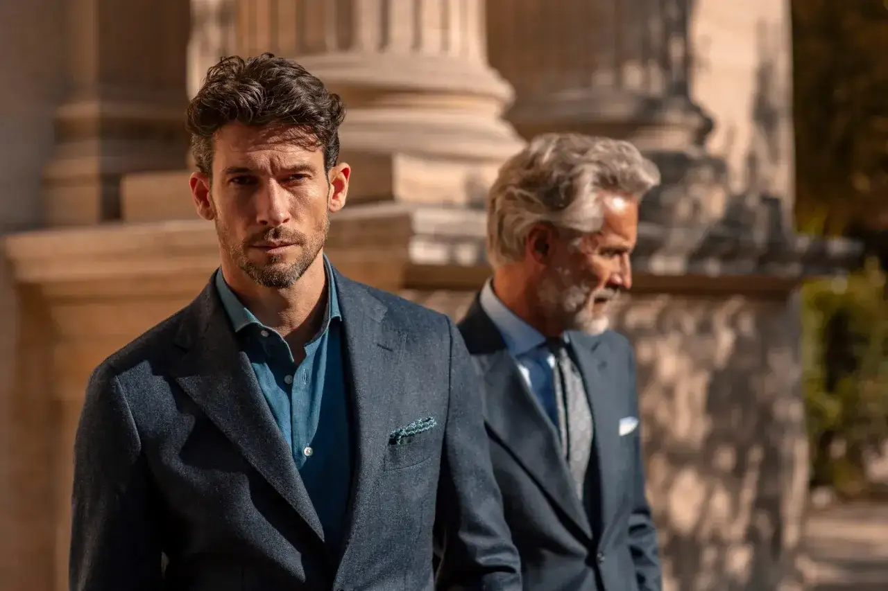 Due uomini eleganti in abito spezzato con cravatta, uno in primo piano con camicia blu, l'altro sfocato sullo sfondo.