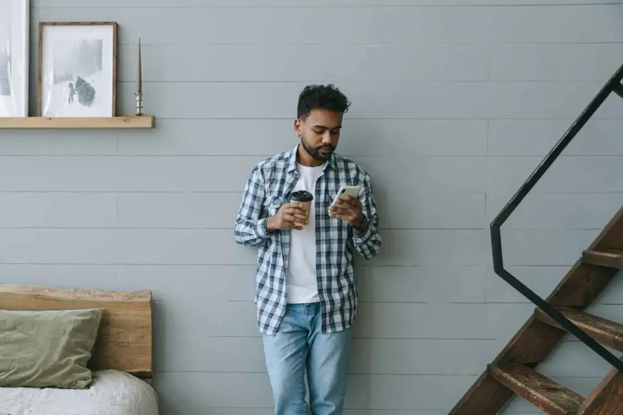 Uomo con camicia a quadri aperta su maglietta bianca, tiene in mano un caff&egrave; e guarda il telefono.