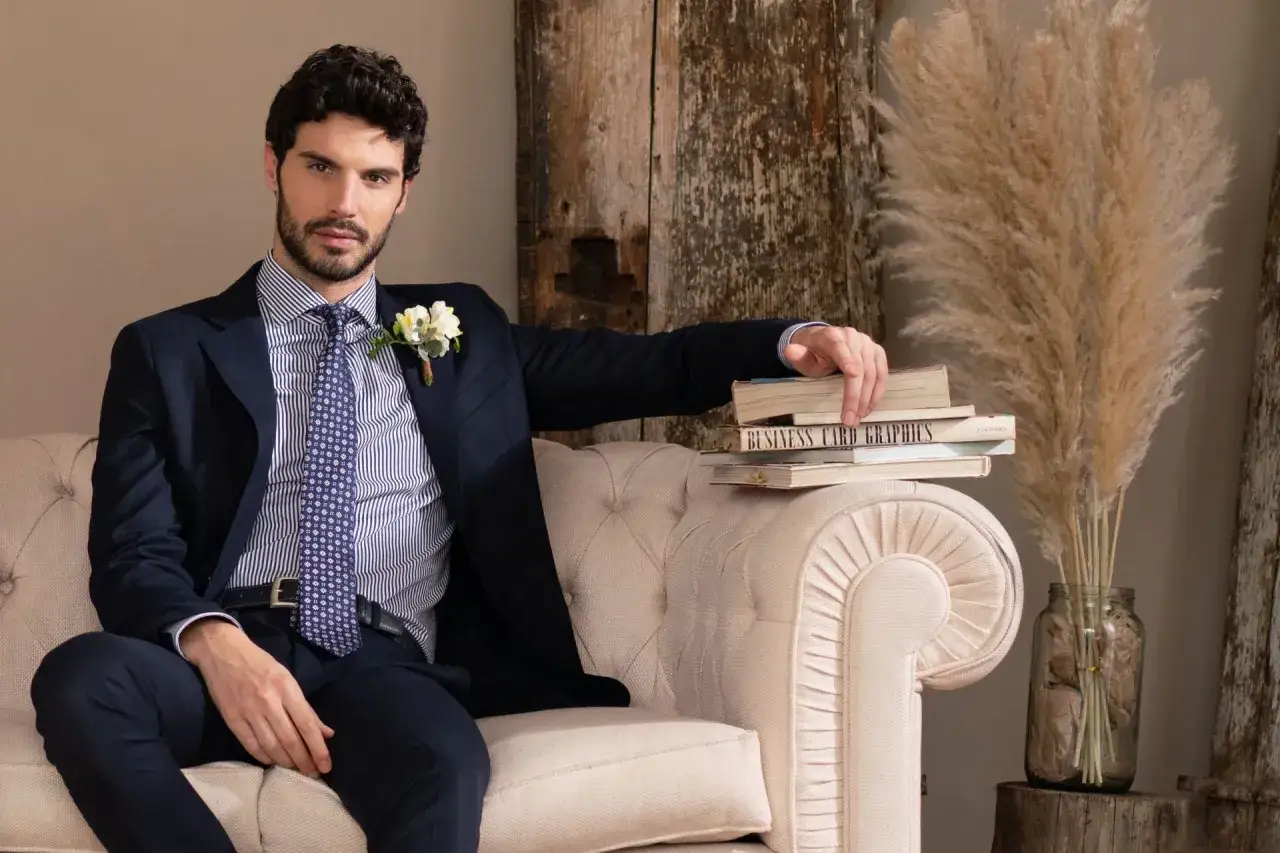 Uomo con camicie da uomo particolari, completo scuro, cravatta a fantasia, seduto su divano, con libri e pampas.