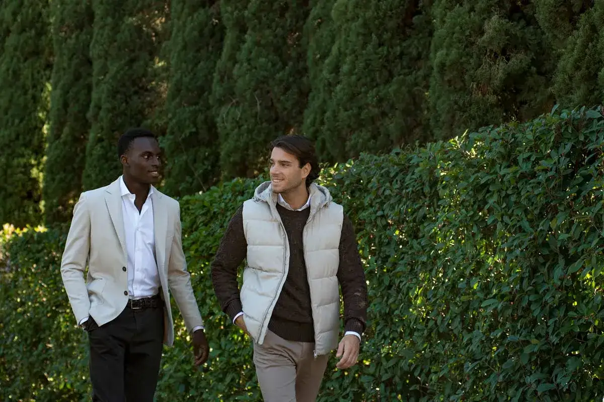 Due uomini passeggiano in un giardino. Uno indossa un completo beige e pantaloni neri, l'altro un gilet panna su un maglione marrone. Il marrone e nero stanno bene insieme, creando un look elegante.