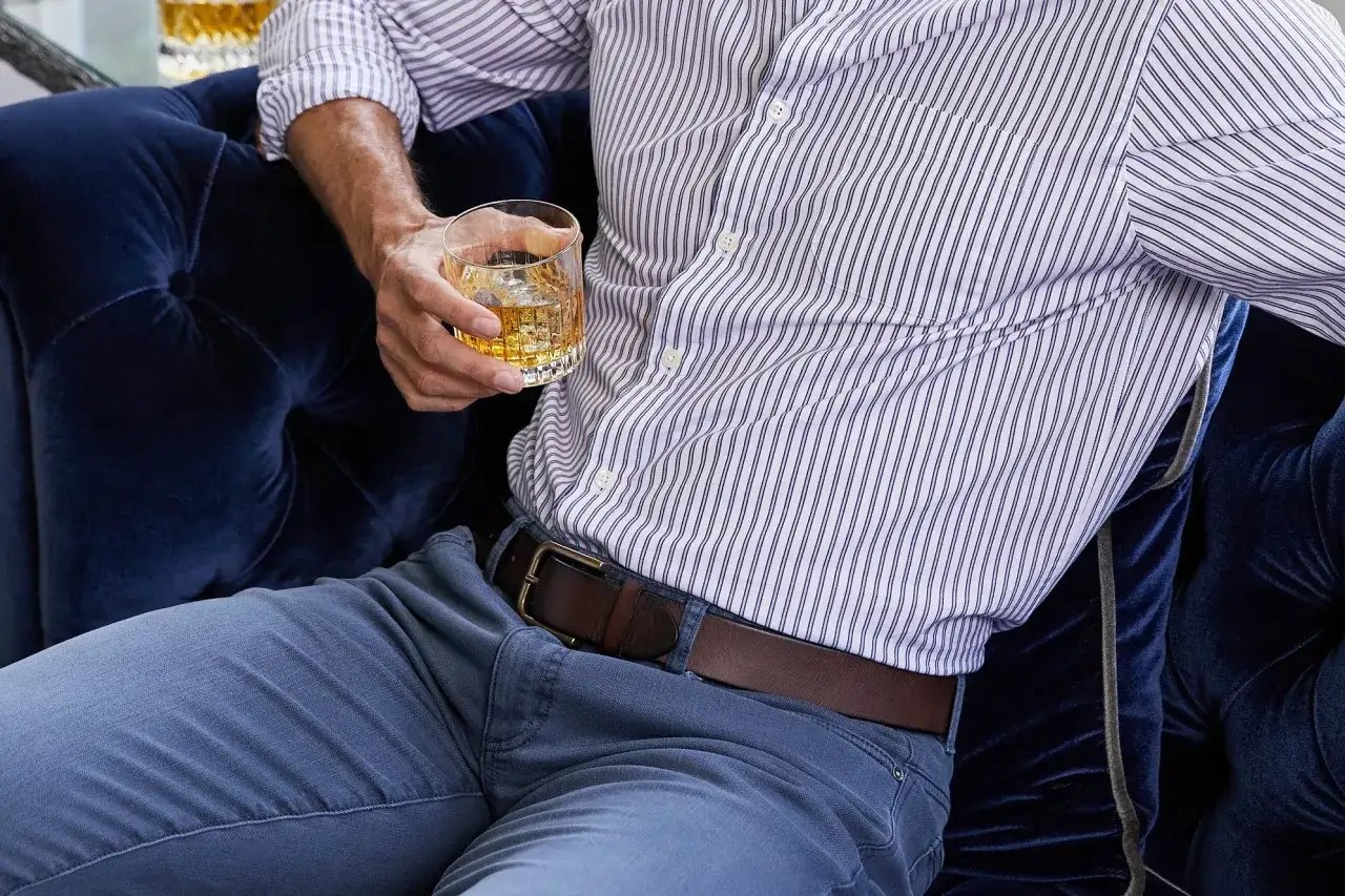 Uomo con camicia a righe e cintura in pelle marrone, tiene un bicchiere di whisky. La cintura uomo &egrave; ben visibile.