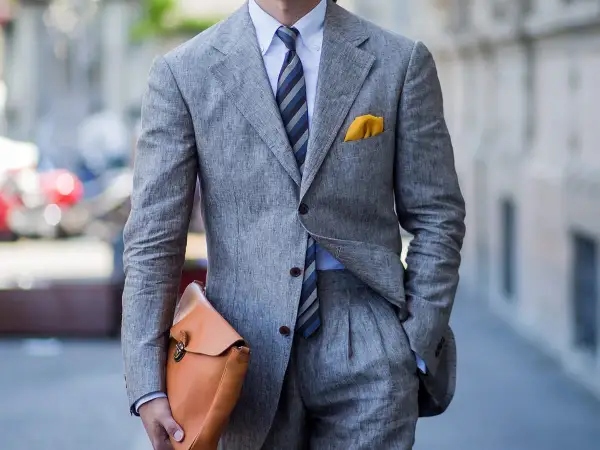Uomo con completo grigio in lino, camicia azzurra e cravatta azzurra a righe. Un perfetto cravatta azzurra abbinamento con pochette gialla.