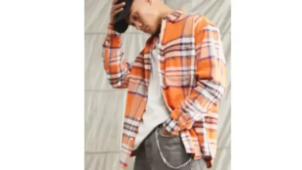 Uomo con camicia in flanella a quadri arancioni, jeans grigi e cappellino nero.