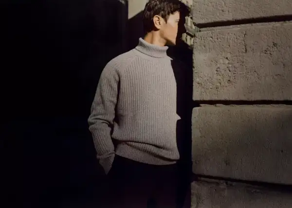 Uomo con un elegante outfit con dolcevita grigio, in piedi vicino a un muro di pietra.