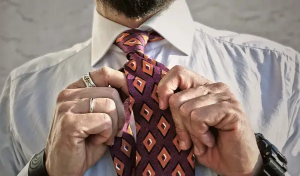 Uomo che sistema il nodo cravatta Eldredge, con camicia bianca e cravatta a rombi arancioni e viola.