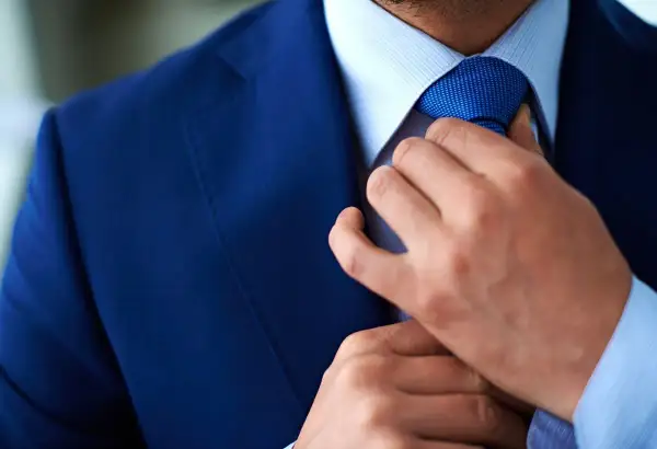 Uomo in abito blu che sistema la cravatta Windsor. Dettaglio elegante e raffinato.