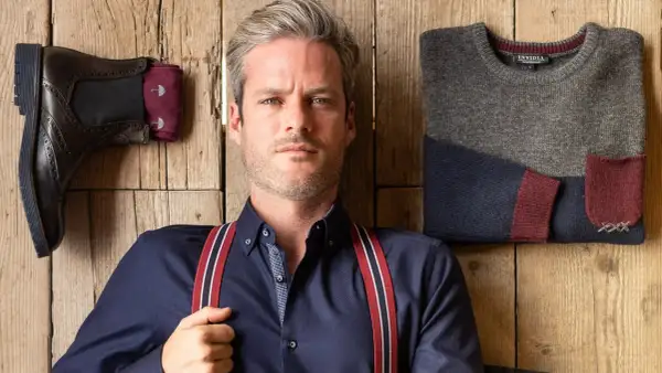Uomo con outfit bretelle, camicia blu, stivali e maglione coordinato su fondo in legno.
