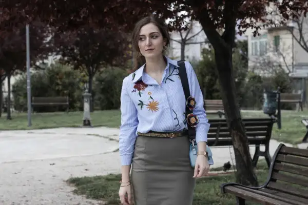 Donna con camicie ricamate a fiori, in un parco con panchine e alberi.