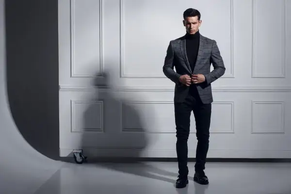 Uomo con maglione nero a collo alto e giacca a quadri grigia, pantaloni scuri e scarpe eleganti.