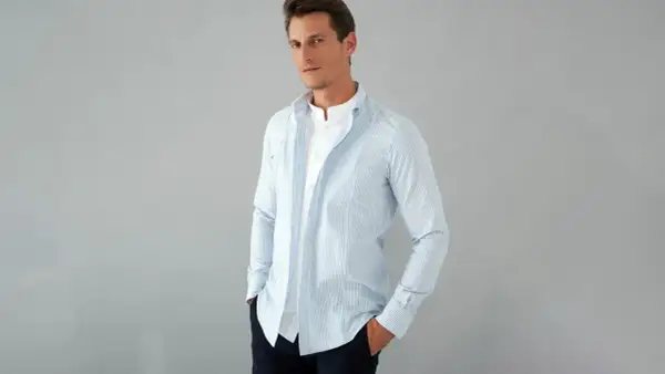 Uomo con outfit camicia a righe azzurre e bianche, indossata aperta su una t-shirt bianca. Mani nelle tasche, look casual.