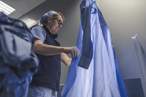 Donna con occhiali e cappellino blu sistema un camice azzurro, forse in una lavanderia.