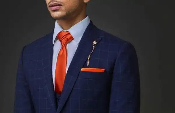 Uomo con abito blu a quadri, camicia azzurra e cravatta arancione con nodi cravatta particolari. Taschino e spilla coordinati.