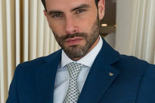 Uomo con vestito blu, camicia bianca e cravatta a fantasia. Sguardo intenso, barba curata.